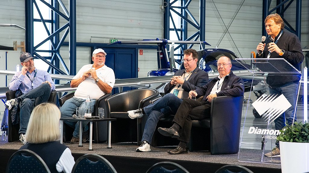 Five men engage in a lively discussion on stage, with one speaker using a microphone while others listen attentively in a hangar setting.