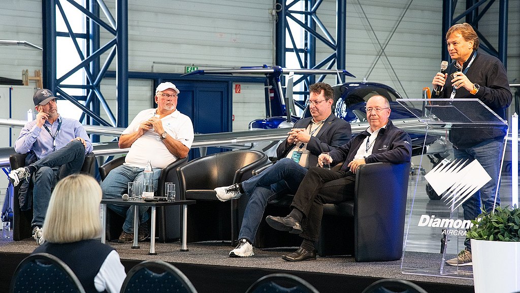 Five men engage in a lively discussion on stage, with one speaker using a microphone while others listen attentively in a hangar setting.