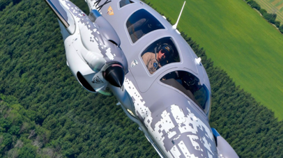 A modern DA62 MPP aircraft flies over lush green fields, showcasing its unique camouflage design and a focused pilot inside.