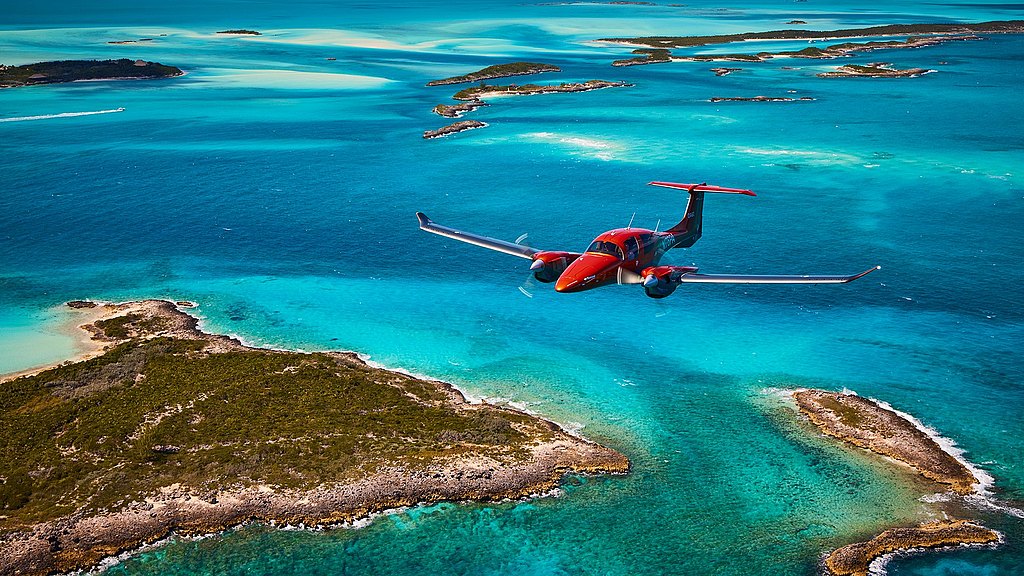 A vibrant red DA62 aircraft soars above turquoise waters, showcasing a stunning view of scattered islands below.