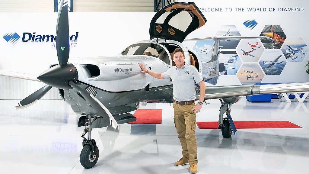 A smiling man poses next to a DA50 RG, with its propeller and open gullwing doors visible, set against a backdrop of aviation imagery.
