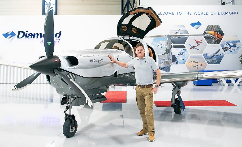 A smiling man poses next to a DA50 RG, with its propeller and open gullwing doors visible, set against a backdrop of aviation imagery.