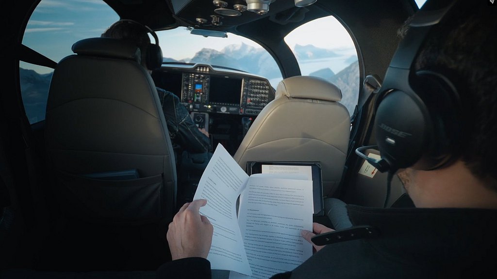 Inside a DA62 aircraft, a passenger reviews documents while the pilot navigates through stunning mountain scenery.