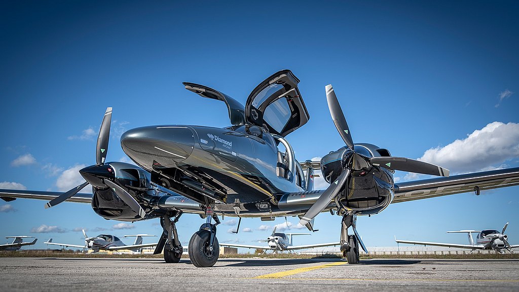 The front view of a stylish DA62 airplane on the tarmac, featuring open gull-wing doors and propellers, with a vibrant sky backdrop.