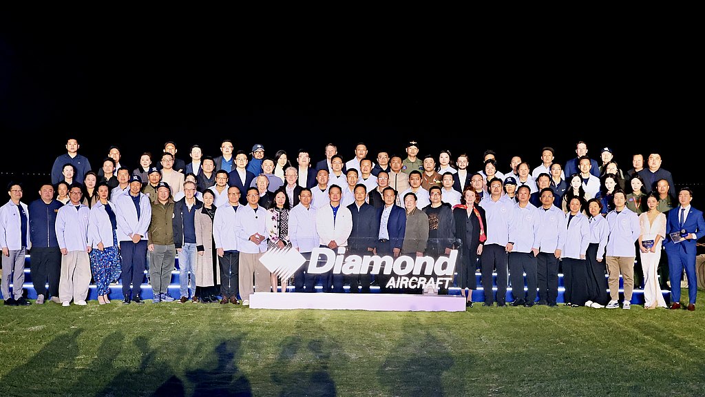A large group of people stands together at night, illuminated by lights, with a prominent 'Diamond Aircraft' sign in front.