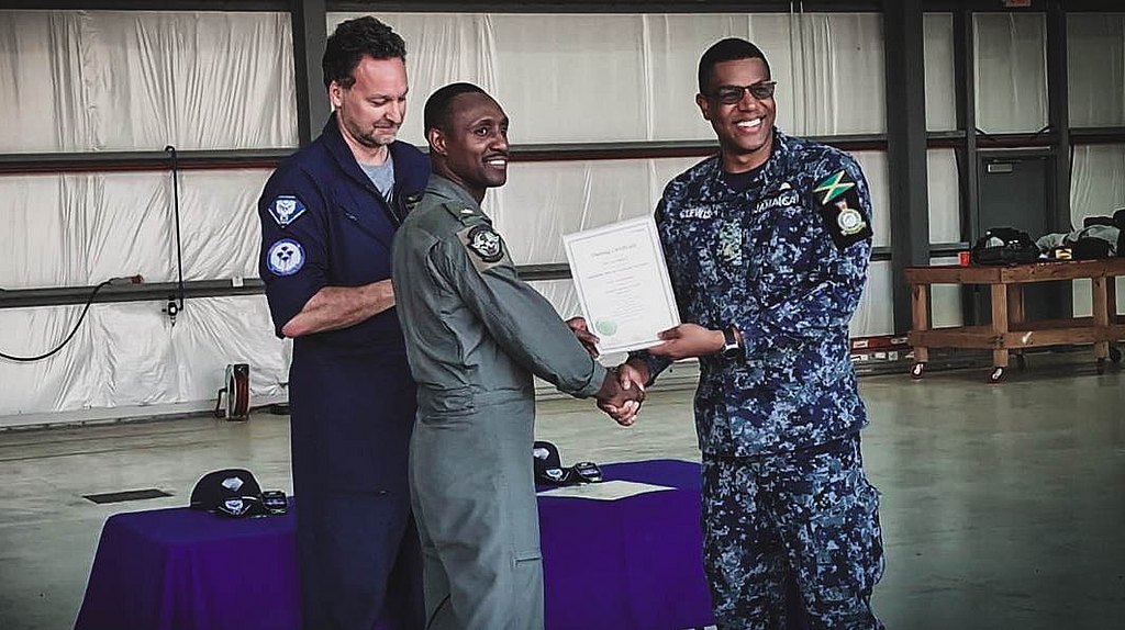 A man in a flight suit shakes hands with a smiling pilot while receiving a certificate in a hangar.