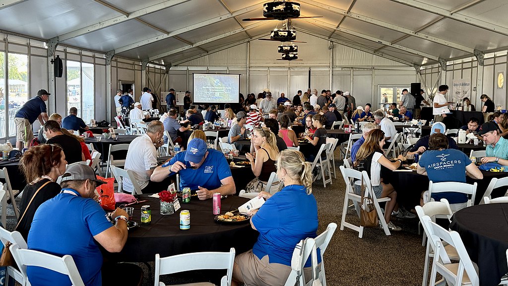 A crowded tent filled with people socializing and eating, featuring a mix of casual attire and a vibrant, festive ambiance.