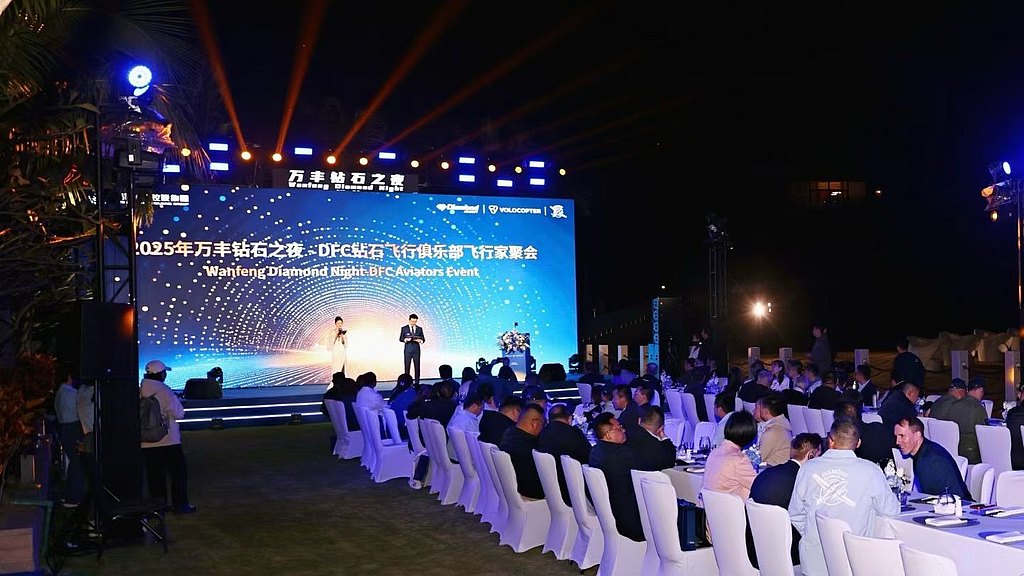An elegant outdoor event features a stage with two presenters, surrounded by guests seated at tables under bright lights.