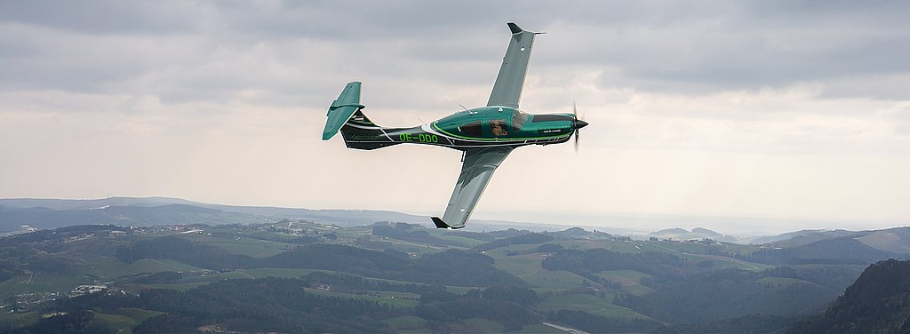 A green DA50 RG aircraft performs a sharp turn in the sky, showcasing its aerodynamic design against a backdrop of rolling hills.