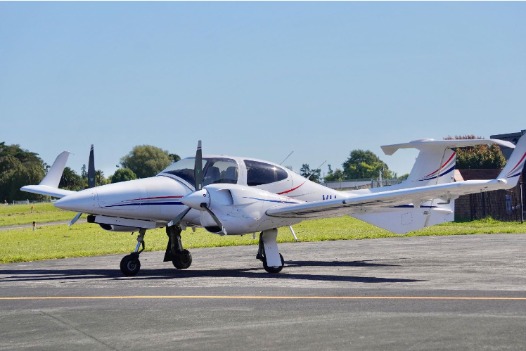 A sleek white DA42-VI aircraft with red and blue stripes is parked on a sunny airstrip, surrounded by green grass and trees.